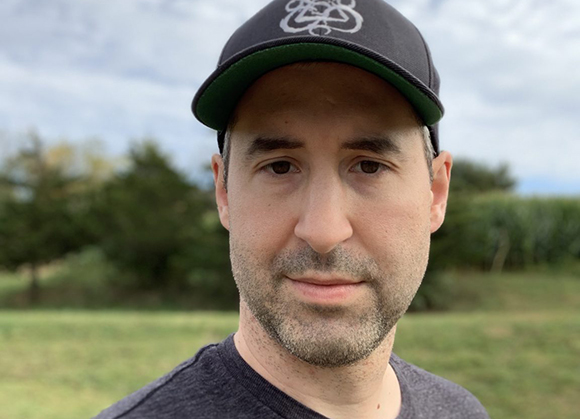 A man with short dark hair and stubble wearing a dark baseball cap and grey t-shirt.