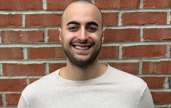 A smiling man with a shaved head and short beard wearing a light beige sweater against a red brick wall.