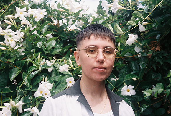 A person with short dark hair and round glasses stands before a lush bush of white flowers.