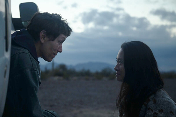 A man and a woman sit facing each other in a dark, open landscape under a cloudy sky.