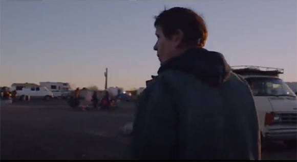 A person in a dark jacket stands in the foreground, looking toward parked vehicles under a clear sky.