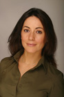 A woman with dark hair and brown eyes wearing an olive green collared shirt against a gray background.