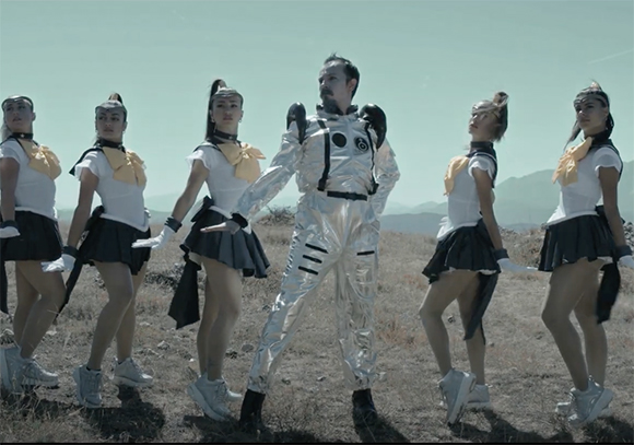 A man in a silver jumpsuit stands among five women in white and black sailor outfits in a dry field.