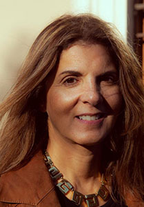 A woman with long brown hair and a patterned necklace smiles at the camera.