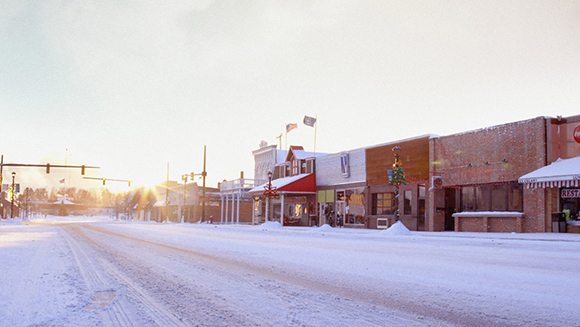 A snow-covered street lined with small storefronts under a bright, hazy sunset sky.