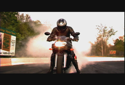 A person in black gear on a motorcycle performing a burnout with thick white smoke rising around them.