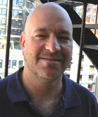 A smiling bald man with light facial hair wearing a dark blue polo shirt outdoors.