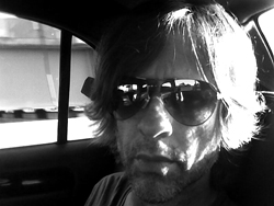 Black and white close-up of a man with medium-length hair wearing sunglasses inside a car.
