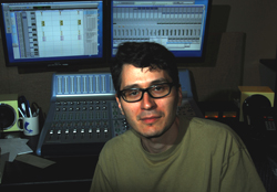 A man with glasses sits in a dark studio before two computer monitors and a large mixing console.