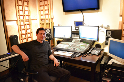 A man sits in a chair at a desk filled with computer monitors, a mixing console, and studio speakers.
