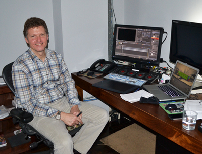 A man in a plaid shirt sits at a wooden desk equipped with two monitors and video editing hardware.