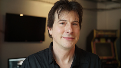 A man with dark hair and a black button-down shirt smiles slightly in a dimly lit room.