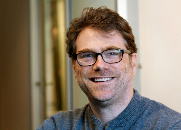 A smiling man with brown hair and black-rimmed glasses wearing a blue sweater.