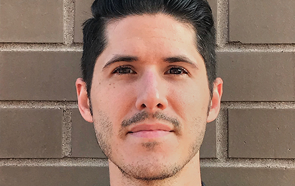 Close-up portrait of a man with dark hair and a light beard looking forward against a brown brick wall.
