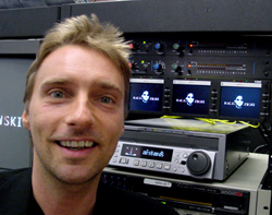 A man smiling in front of a rack of professional audio recording equipment and monitors.