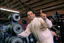 A man in a light sweatshirt points his index finger up in a gym filled with weightlifting equipment.