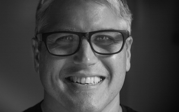 Black and white close-up portrait of a man with short hair and glasses smiling.