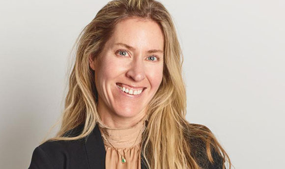 A smiling woman with long blonde hair wearing a black blazer and a tan high-neck top.
