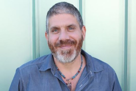 A smiling man with graying hair, a beard, and a beaded necklace wearing a blue button-down shirt.