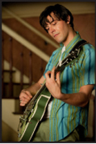 A man in a blue striped shirt playing a green electric guitar indoors.