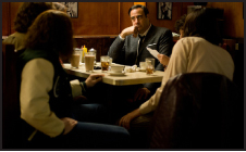 A man in a suit sits at a diner table, looking thoughtful while surrounded by people in a dimly lit booth.
