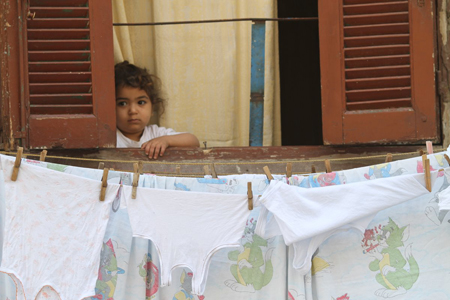 A young child peeks out from a window behind a clothesline of white undergarments.