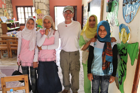 A man in a white shirt and glasses stands with four girls wearing hijabs in a classroom.