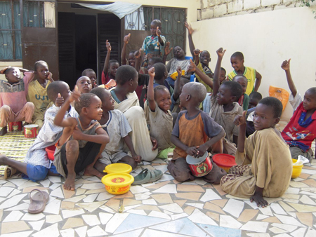 A large group of children sitting and standing on a tiled floor, some raising their hands.