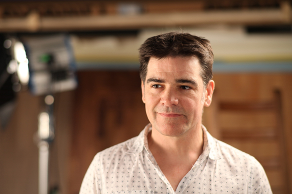 A man with dark hair wearing a white patterned button-down shirt looks to the side.