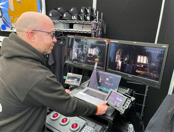 A man in a dark hoodie operates a video editing suite with two large monitors and a laptop.