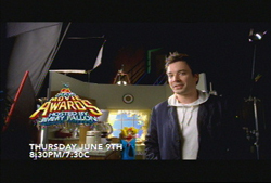 A man in a dark jacket and white hoodie stands in a studio with an MTV Movie Awards graphic.