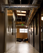 A long hallway with exposed ceiling pipes and metal trays leading to a room with a window and seating.