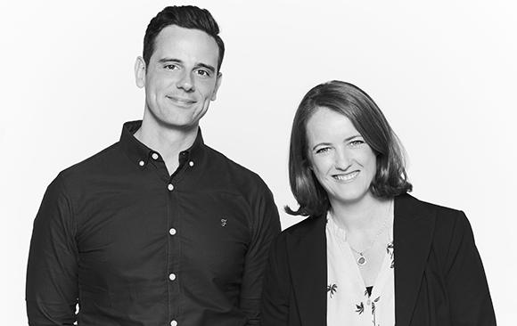 A man in a dark button-down shirt and a woman in a dark blazer and patterned top smile against a white background.
