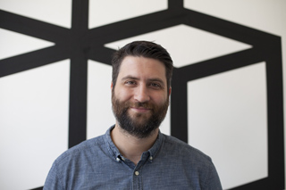 A man with a dark beard and short hair wearing a blue button-down shirt, smiling against a black and white wall.