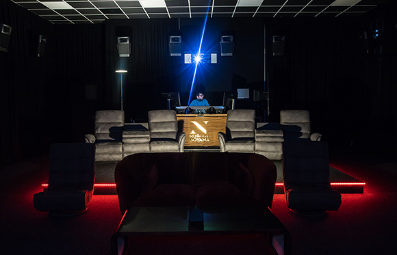 A dark home theater with a large screen, a person at a console, and rows of gray and red seating.