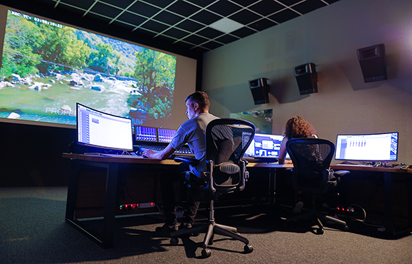 Two people sit at desks with large monitors in a dark studio with a large projection screen.