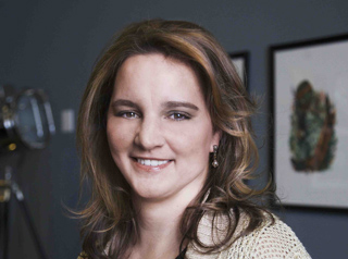 A smiling woman with wavy brown hair and small drop earrings looks toward the camera.