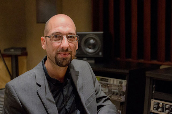 A bald man with glasses and a goatee wearing a grey blazer in a music studio.