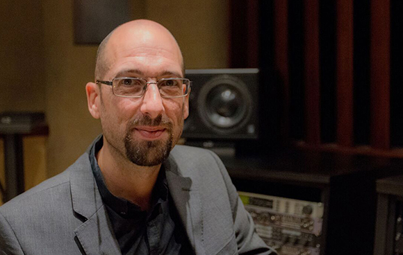 A bald man with glasses and a goatee wearing a grey blazer in a recording studio.