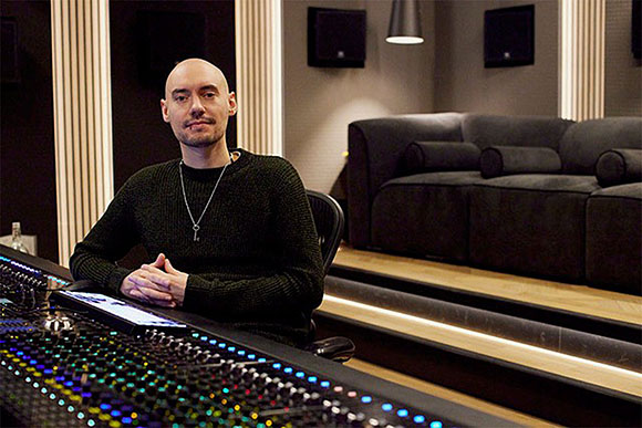 A bald man in a dark sweater sits at a professional audio mixing desk in a studio.