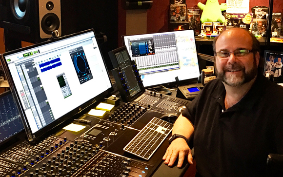 A man with glasses and a beard sits at a large mixing console with multiple computer monitors.