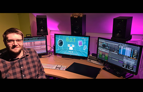 A man in a plaid shirt sits before a desk with three monitors and black studio speakers.