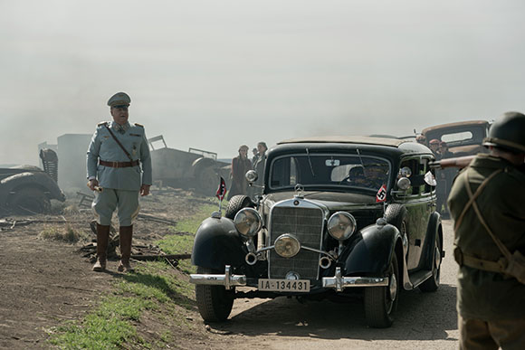 A man in a light grey military uniform stands beside a vintage black car on a dusty road.