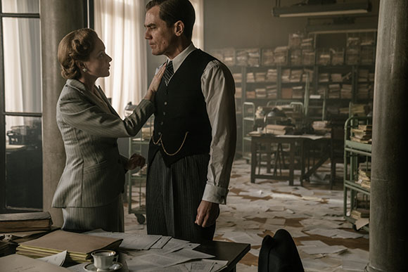 A woman in a grey suit touches the chest of a man in a black vest in a messy, paper-filled office.