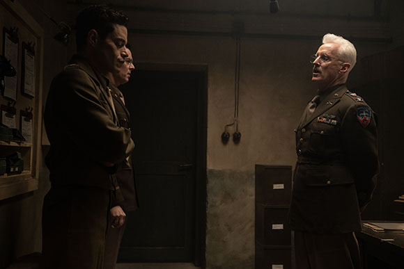 An older man in a military uniform speaks to two other men in uniform in a dark, indoor setting.