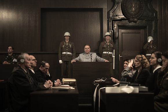 A man in a light grey uniform stands at a podium in a dark courtroom with guards and onlookers.