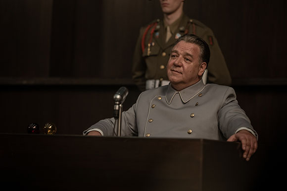 A man in a grey military uniform sits behind a dark wooden podium with a microphone.