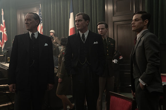 Four men in vintage suits and military uniforms stand in a dimly lit room with flags in the background.