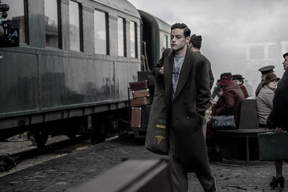 A man in a dark overcoat walks along a train platform next to a passenger train.