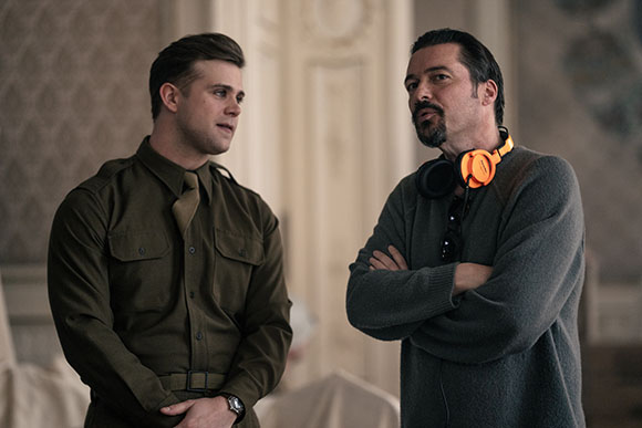A man in a green military-style shirt looks at a man in a grey sweater with orange headphones around his neck.